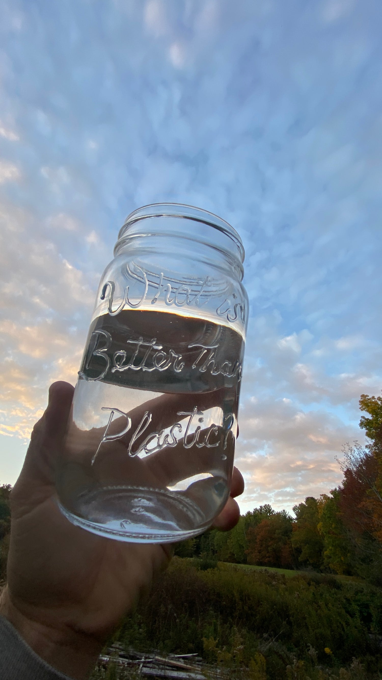 Sunset sky background, with a hand holding up an embossed glass storage mason jar with “What is Better Than Plastic?” Brand Logo